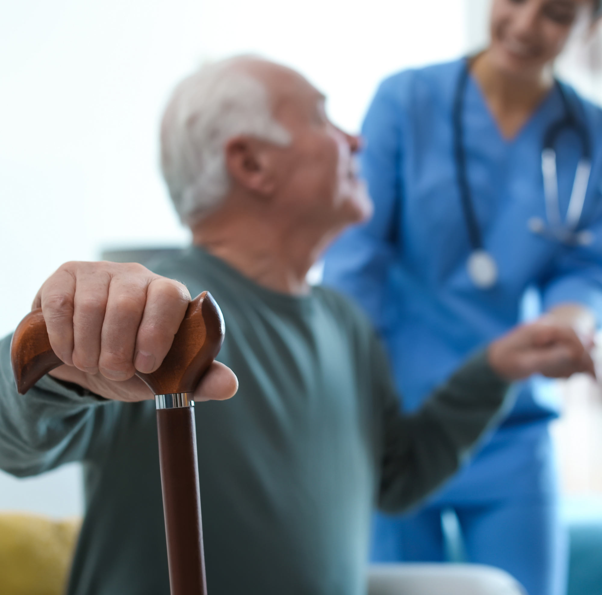 Senior mit Gehstock wird von einer Pflegekraft im blauen Kittel unterstützt.