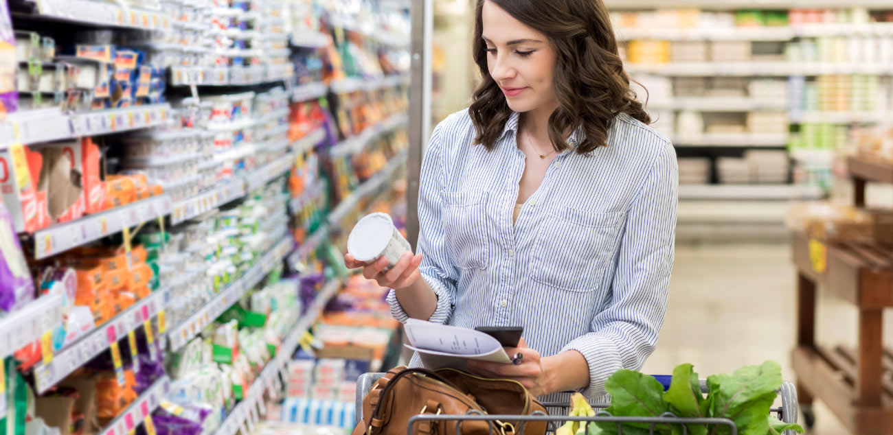 einkaufsservice-supermarkt Frau prüft ein Produkt im Supermarkt und hält dabei eine Einkaufsliste in der Hand.