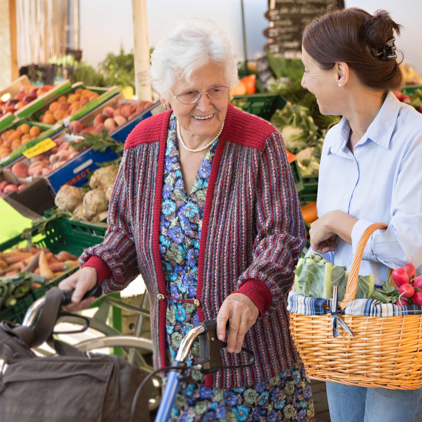 Ältere Frau mit Rollator wird auf einem Markt von einer Begleitperson beim Einkauf unterstützt.