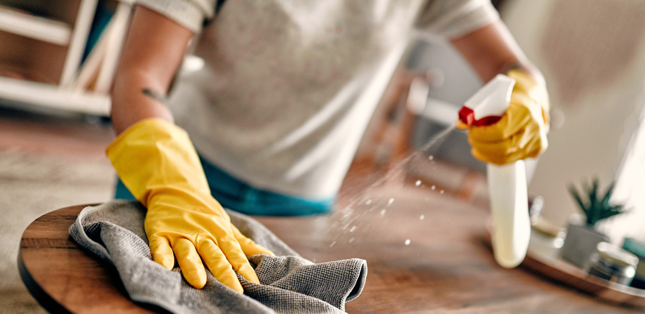 reinigung-der-wohnung Person mit gelben Gummihandschuhen reinigt einen Holztisch mit Spray und Tuch.